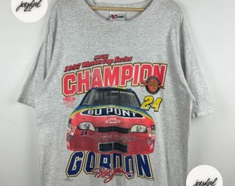 May include: A light gray t-shirt featuring a colorful graphic of a race car with the text "CHAMPION" and "GORDON". The shirt commemorates the 1997 Winston Cup Series.