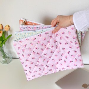 May include: A set of three quilted fabric pouches with floral patterns. The pouches are pink, green, and peach with white floral designs. The pouches are stacked on top of each other and are being held by a hand.