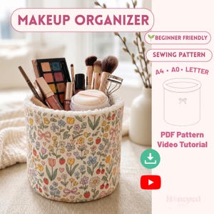 Könnte beinhalten: Ein zylindrischer Make-up-Organizer aus Stoff mit cremefarbenem Hintergrund und einem bunten Blumen-, Erdbeer- und Schleifenmuster. Das Innere ist mit weißem Fleece gefüttert. Er enthält verschiedene Make-up-Pinsel, Paletten und Kosmetika.