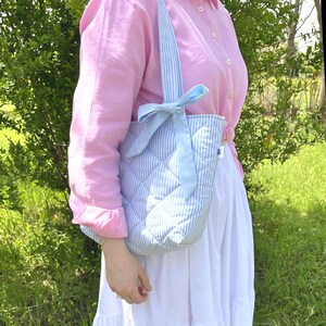 Puede incluir: Un bolso tote acolchado de rayas azules y blancas con un lazo azul claro. El bolso tiene un dise&ntilde;o acolchado blanco y lo lleva una persona que lleva una camisa rosa y una falda blanca.