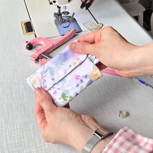 May include: A small rectangular fabric pouch featuring a watercolour floral print in shades of purple, blue, yellow, and green. The pouch has a flap closure secured with a white plastic snap button, positioned near a pink handheld snap fastener tool.
