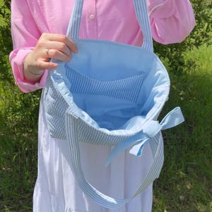 Puede incluir: Una bolsa de mano de rayas azules y blancas con un lazo en el asa. La bolsa est&aacute; abierta y el interior est&aacute; forrado con un tejido azul claro.