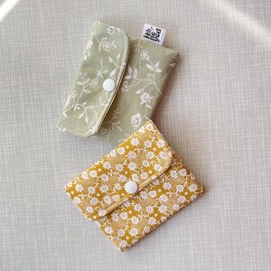 May include: Two small fabric pouches with snap closures. One pouch is sage green with a white floral vine pattern, and the other is mustard yellow with a dense white floral print. Each features a white plastic snap and a small white brand tag.