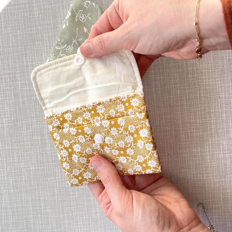 May include: A small fabric pouch featuring a mustard yellow exterior with a white floral pattern and a cream-coloured interior lining. The pouch has a fold-over flap secured by a single white plastic snap button, revealing a sage green floral fabric.