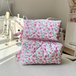 K&ouml;nnte beinhalten: Zwei rechteckige, gesteppte Stofftaschen, die vertikal gestapelt sind und einen wei&szlig;en Hintergrund mit einem sich wiederholenden Muster aus rosa Aquarellrosen und gr&uuml;nen Bl&auml;ttern aufweisen. Jede Tasche hat einen seitlichen Rei&szlig;verschluss.