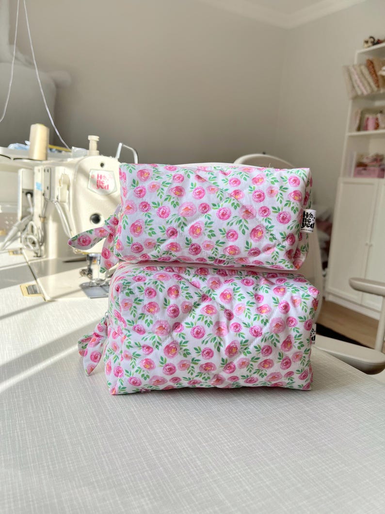 K&ouml;nnte beinhalten: Zwei rechteckige, gesteppte Stofftaschen, die vertikal gestapelt sind und einen wei&szlig;en Hintergrund mit einem sich wiederholenden Muster aus rosa Aquarellrosen und gr&uuml;nen Bl&auml;ttern aufweisen. Jede Tasche hat einen seitlichen Rei&szlig;verschluss.