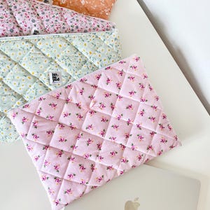 May include: Three quilted fabric laptop sleeves with floral patterns. The sleeves are in shades of pink, green, and brown. The text "He + She" is visible on the green sleeve.