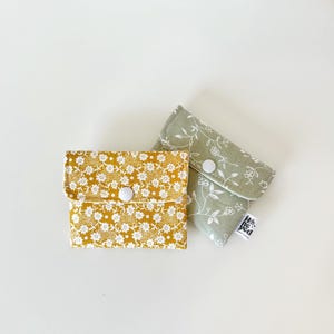 May include: Two small fabric pouches with snap closures. One pouch is mustard yellow with a dense white floral pattern. The other is sage green with a sparse white vine and flower print. A small white tag with the text "honeyed" is attached.