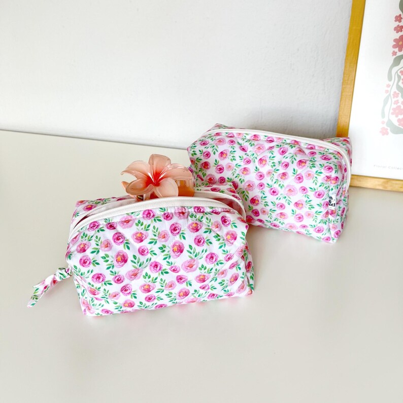 K&ouml;nnte beinhalten: Zwei rechteckige Rei&szlig;verschlusstaschen mit wei&szlig;em Hintergrund und einem wiederkehrenden Muster aus rosa Rosen und gr&uuml;nen Bl&auml;ttern. Auf einer Tasche liegt eine hellrosa Blume auf dem geschlossenen wei&szlig;en Rei&szlig;verschluss. Beide haben eine Steppoptik.
