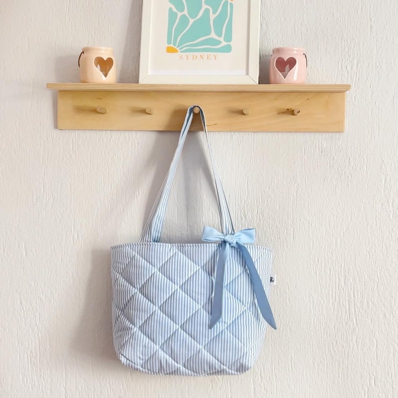 Puede incluir: Un bolso tote acolchado azul claro con rayas blancas y un lazo azul claro. El bolso est&aacute; colgado en una estanter&iacute;a de madera con ganchos.