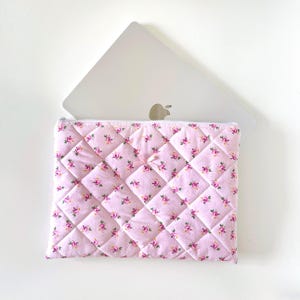 May include: A pink quilted laptop sleeve with a floral pattern. The sleeve has a zipper closure and a small bow on the front.