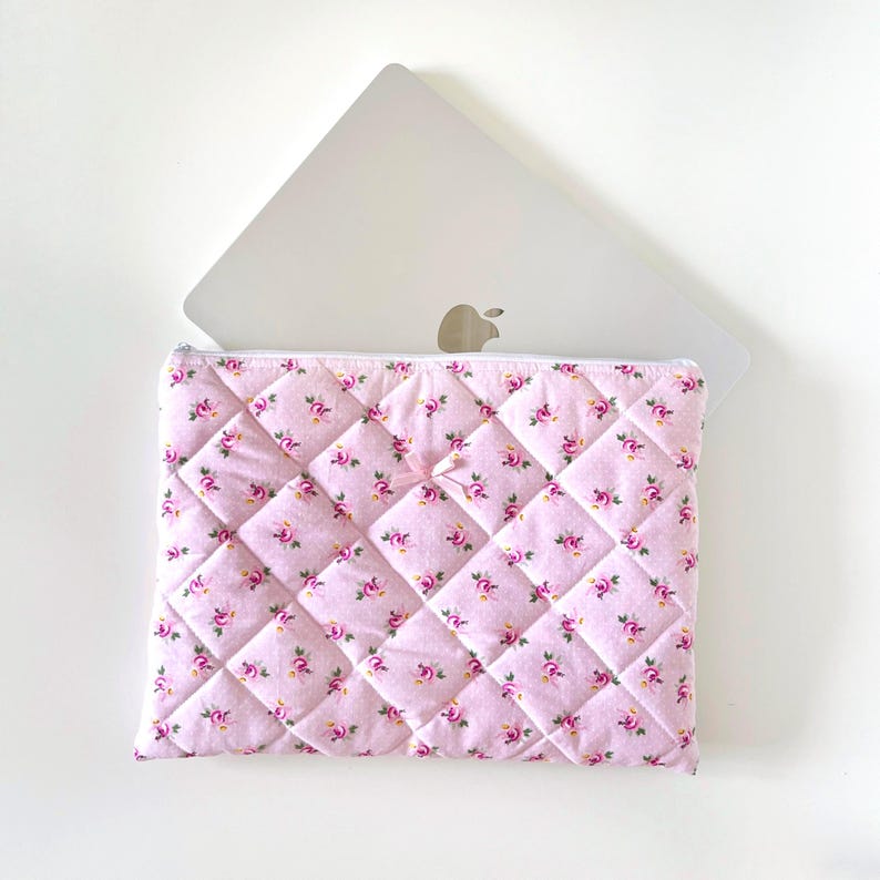 May include: A pink quilted laptop sleeve with a floral pattern. The sleeve has a zipper closure and a small bow on the front.