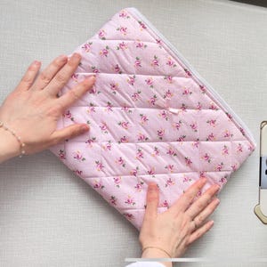 May include: A pink quilted zippered pouch with a floral pattern. The pouch is folded in half and held by two hands.