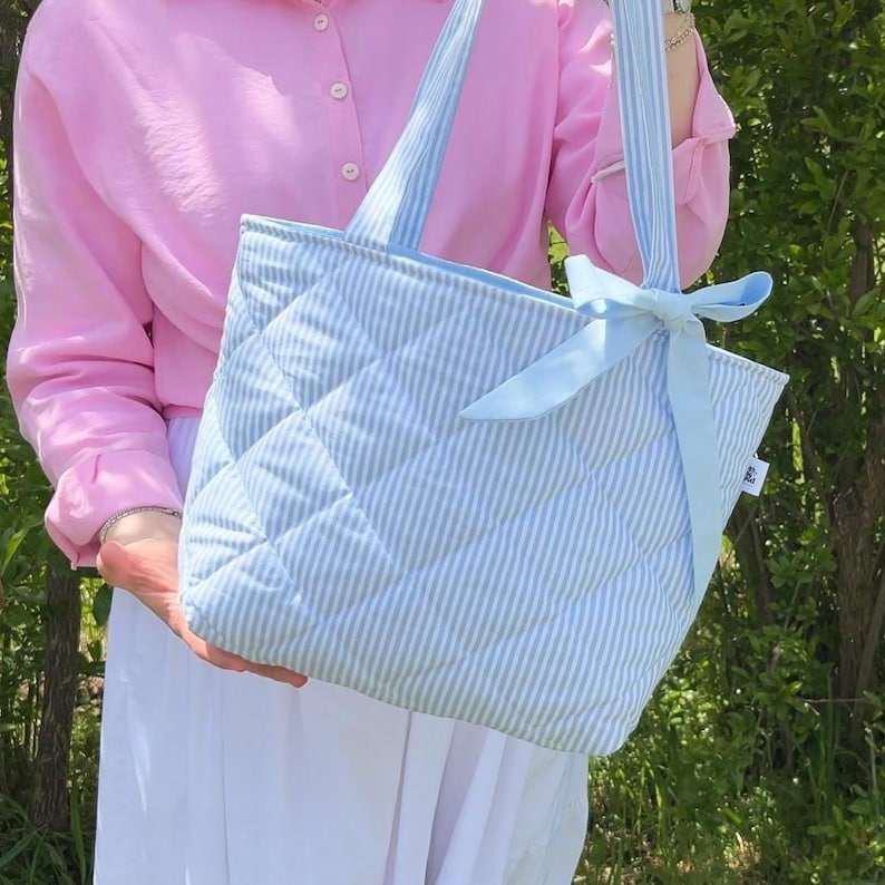 Puede incluir: Una bolsa de mano acolchada de color azul claro con rayas blancas y un lazo de cinta a juego. La bolsa tiene dos asas y un dise&ntilde;o acolchado.