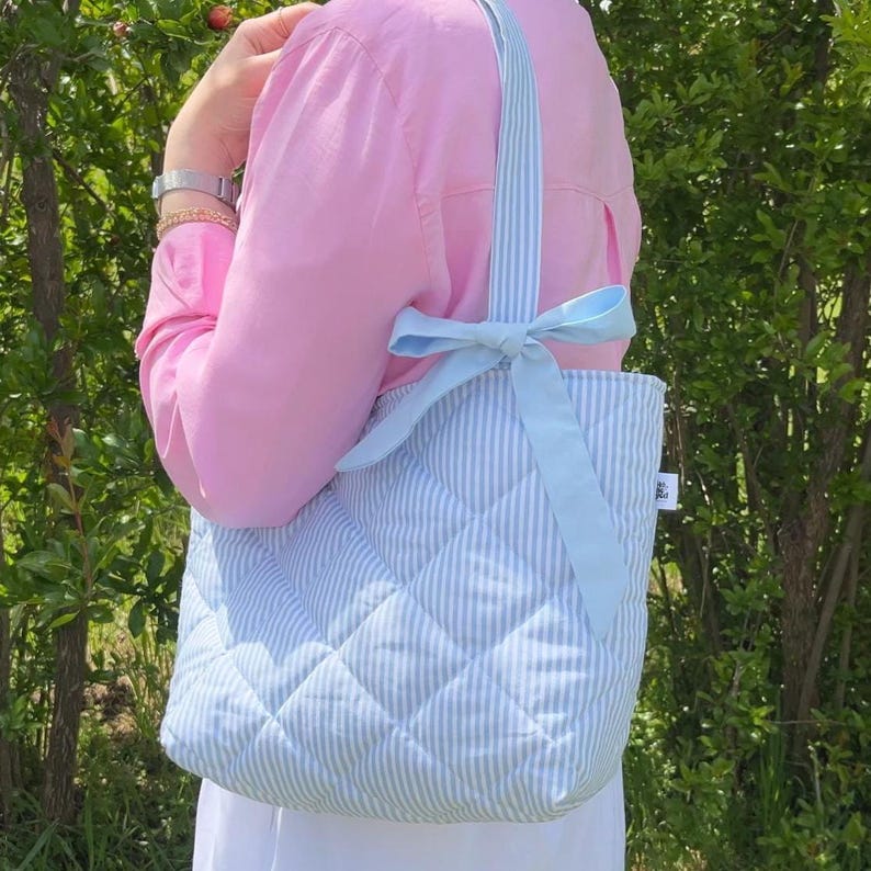 Puede incluir: Un bolso tote acolchado con rayas azules y blancas. El bolso tiene un lazo azul claro y una correa blanca.