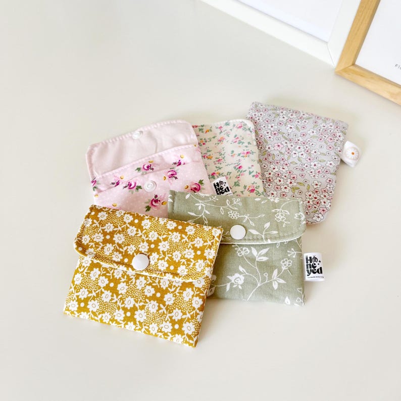 May include: A collection of four small fabric pouches with snap closures, featuring various floral patterns in mustard yellow, sage green, light pink, and grey. Each pouch has a small white tag with the text "Honeyd" printed in black.