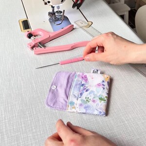 May include: A small, rectangular fabric pouch featuring a floral pattern in shades of purple, blue, and green on one side and solid light purple on the other. The pouch has a white snap fastener and a small fabric tag with text on the edge.