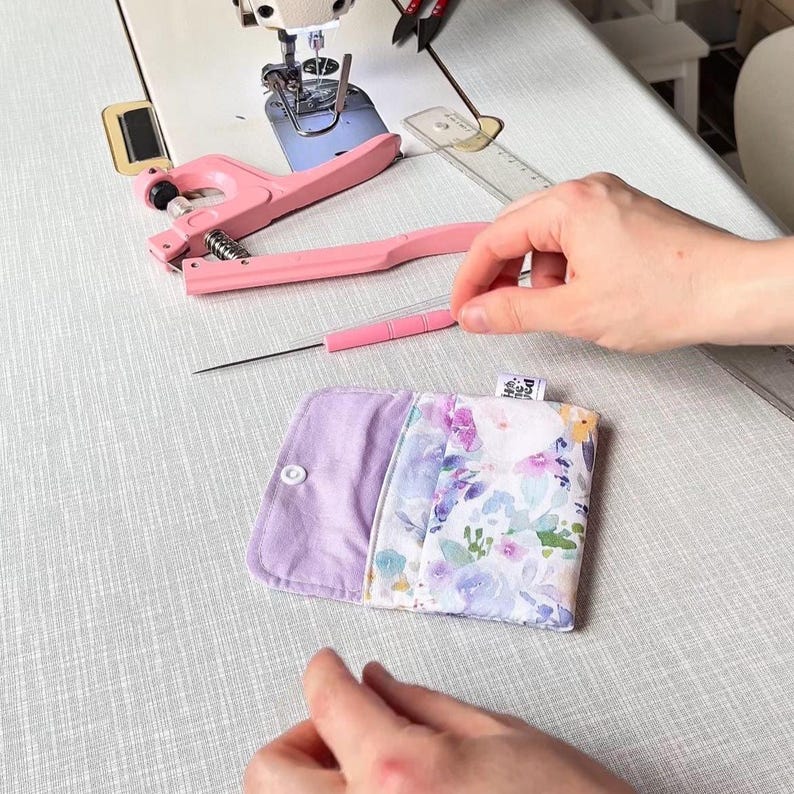 May include: A small, rectangular fabric pouch featuring a floral pattern in shades of purple, blue, and green on one side and solid light purple on the other. The pouch has a white snap fastener and a small fabric tag with text on the edge.