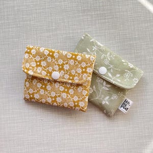 May include: Two small fabric pouches with snap closures. One pouch is mustard yellow with a dense white floral pattern, and the other is sage green with a sparse white line-art floral design. A small white tag reads honeyed on the green pouch.