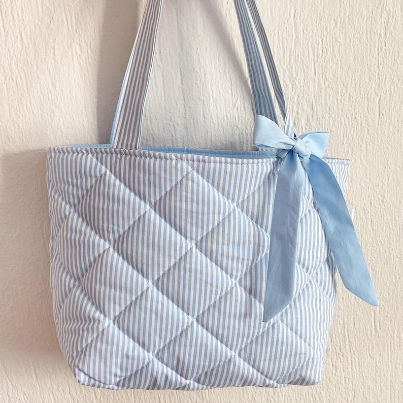 Puede incluir: Una bolsa de mano acolchada con rayas azules y blancas y un lazo de sat&eacute;n azul. La bolsa tiene un patr&oacute;n acolchado de rombos.