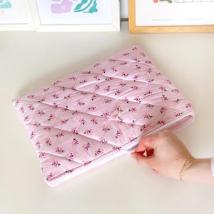 May include: A pink quilted zippered pouch with a floral pattern. The zipper is open and a hand is reaching in.