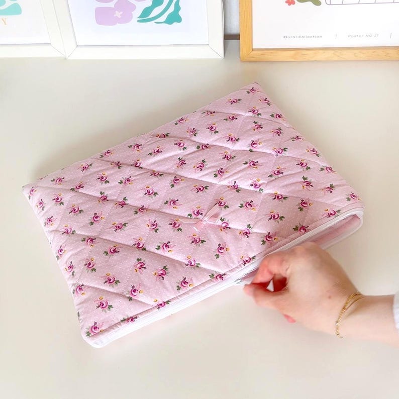 May include: A pink quilted zippered pouch with a floral pattern. The zipper is open and a hand is reaching in.