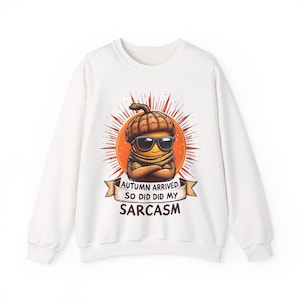 Könnte beinhalten: Ein weißes Langarm-Sweatshirt mit einer Grafik einer Eichel, die eine Sonnenbrille und einen Schal trägt, vor einer orangefarbenen Sonne. Unter der Eichel befindet sich ein Banner mit dem Text: AUTUMN ARRIVED. SO DID DID MY SARCASM.