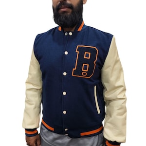 Op de afbeelding: Een marineblauw varsity-jack met crèmekleurige mouwen, voorzien van een grote oranje letter B-patch op de borst. Het jack heeft een sluiting met drukknopen, geribbelde boorden met oranje strepen en een bijpassende geribbelde kraag en zoom.