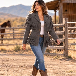 May include: A charcoal gray suede jacket featuring long fringe detailing across the chest and hem, a collared neckline, and a button-front closure. The jacket is worn over a white top with blue denim jeans and brown leather cowboy boots.