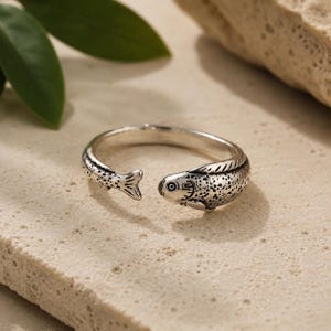 Puede incluir: Un anillo abierto de tono plateado con forma de pez. La banda presenta una superficie texturizada y detallada con marcas oscuras y moteadas en el cuerpo, una cabeza definida con un ojo y una aleta caudal en el extremo opuesto del diseño.