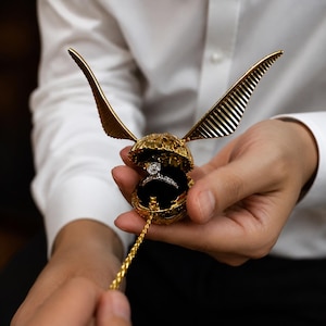 Puede incluir: Un joyero esférico de color dorado con dos alas extendidas, diseñado para parecer una bola voladora. El joyero está abierto y muestra un anillo de tono plateado con una piedra redonda central sobre un forro de terciopelo negro.