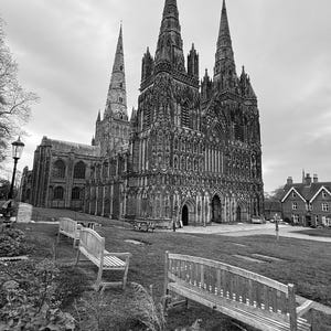 May include: A black and white photograph of a large, gothic cathedral with two tall spires. The cathedral is surrounded by a grassy lawn with two wooden benches in the foreground.