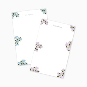 May include: Two white notepads with floral corner designs. One notepad has blue flowers and the text "ON MY LIST". The other notepad has purple flowers and the text "LILY NEWTON".