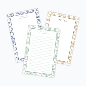 May include: Three notepads with floral borders in blue, green, and gold. The blue notepad has the text "from the desk of EMILY LAURENT". The green notepad has lined paper and the text "from the desk of STEVIE MITCHELL". The gold notepad has the text "NOTES".