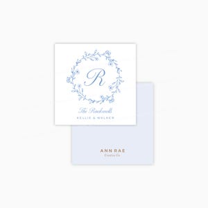 May include: Square white card with a blue floral wreath design around the letter "R". The text "The Rockwells" and "Kellie & Walker" are below the wreath. A light blue envelope is beneath the card. The text "ANN RAE Creative Co." is on the envelope.