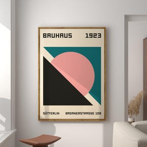 Puede incluir: Un póster Bauhaus enmarcado de 1923. El diseño presenta un círculo rosa, un triángulo negro y rectángulos turquesa y crema. El póster incluye la palabra "BAUHAUS" en la parte superior y el texto "SÜTTERLIN BADNERSTRASSE 109" en la parte inferior.