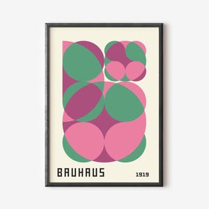 May include: A framed art print with a Bauhaus design. It features overlapping shapes in pink, green, and purple. The word "BAUHAUS" is printed in black at the bottom, with "1919" next to it. The artwork is set against a cream background, with a black frame.