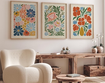 May include: Three framed floral art prints. The prints feature colorful flowers in shades of blue, orange, red, and pink. The frames are a light wood colour. The art is displayed above a wooden console table with a white armchair in the foreground.