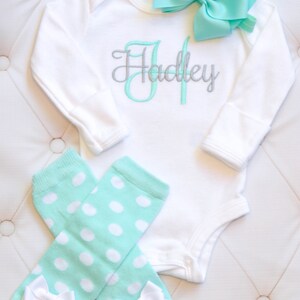 May include: A white long-sleeve bodysuit with the name "Hadley" embroidered in silver and mint green. A mint green headband with a bow is included. The set also includes a pair of mint green leg warmers with white polka dots and white bows.