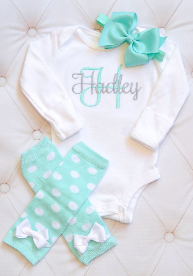 May include: A white long-sleeve bodysuit with the name "Hadley" embroidered in silver and mint green. A mint green headband with a bow is included. The set also includes a pair of mint green leg warmers with white polka dots and white bows.