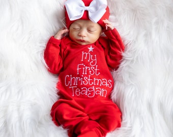 May include: A newborn baby wearing a red Christmas outfit and hat with a large white bow. The outfit has the text "My First Christmas Teagan" embroidered in white. The baby is lying on a fluffy white surface.