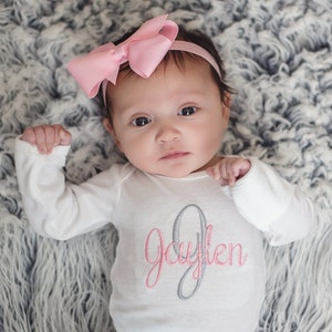 May include: A baby wearing a white long-sleeved onesie with the name "Jaylen" embroidered in pink and gray. The baby is wearing a pink bow headband and is lying on a gray fluffy blanket.