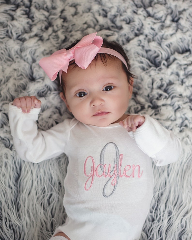 May include: A baby wearing a white long-sleeved onesie with the name "Jaylen" embroidered in pink and gray. The baby is wearing a pink bow headband and is lying on a gray fluffy blanket.