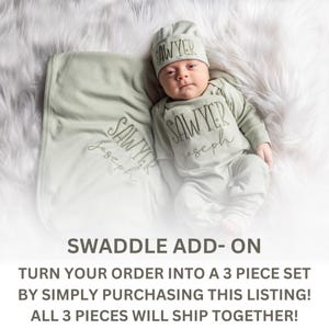May include: A baby wearing a sage green outfit, including a hat, onesie, and pants, with the name "SAWYER" printed on each piece. A matching blanket with the name is also present. The text "SWADDLE ADD-ON" is visible.
