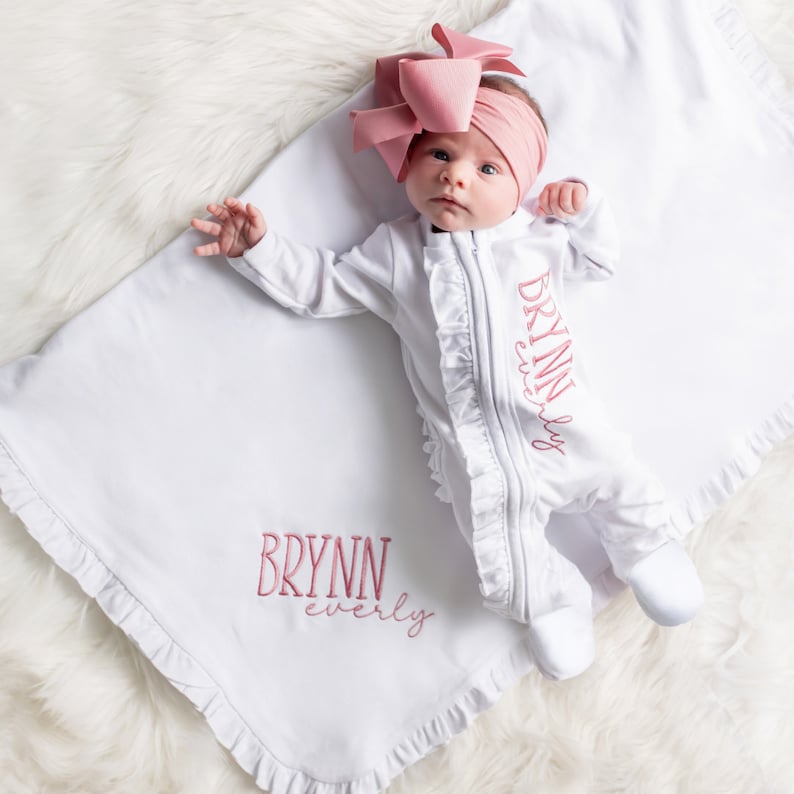 perfect gift for baby girl or baby girl coming home outfit.  A white baby onesie with pink ruffled trim and the text 'BRYNN everly' embroidered on the front. The onesie is lying on a white blanket with a ruffled edge and the text 'BRYNN everly' embroidered on the blanket.