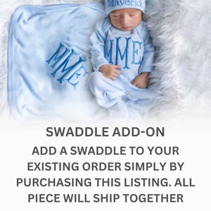 May include: A light blue baby swaddle with the monogram "MME" embroidered on it. The swaddle is lying on a white fluffy surface.