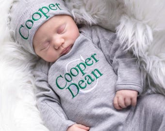 May include: A gray baby onesie with green embroidery that reads "Cooper & Dean". A gray knit hat with green embroidery that reads "Cooper" is on the baby's head.