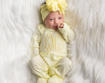 May include: A baby wearing a yellow long-sleeved onesie with a monogram embroidered on the front. The onesie has matching yellow footie socks. The baby is wearing a yellow bow headband.