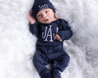 May include: Navy blue baby outfit set with a matching knotted hat. The hat is embroidered with the name "Jett" in white. The onesie features the embroidered initials "JAJ" in white. The baby is lying on a white fluffy surface.