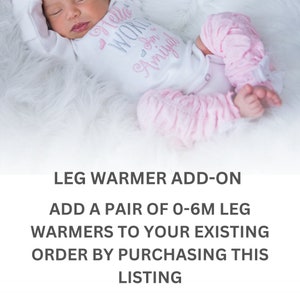 May include: A newborn baby wearing a white onesie with the text "Hello World" and pink leg warmers. The leg warmers have a ruffled design and are paired with a white onesie. The baby is lying on a fluffy white surface.